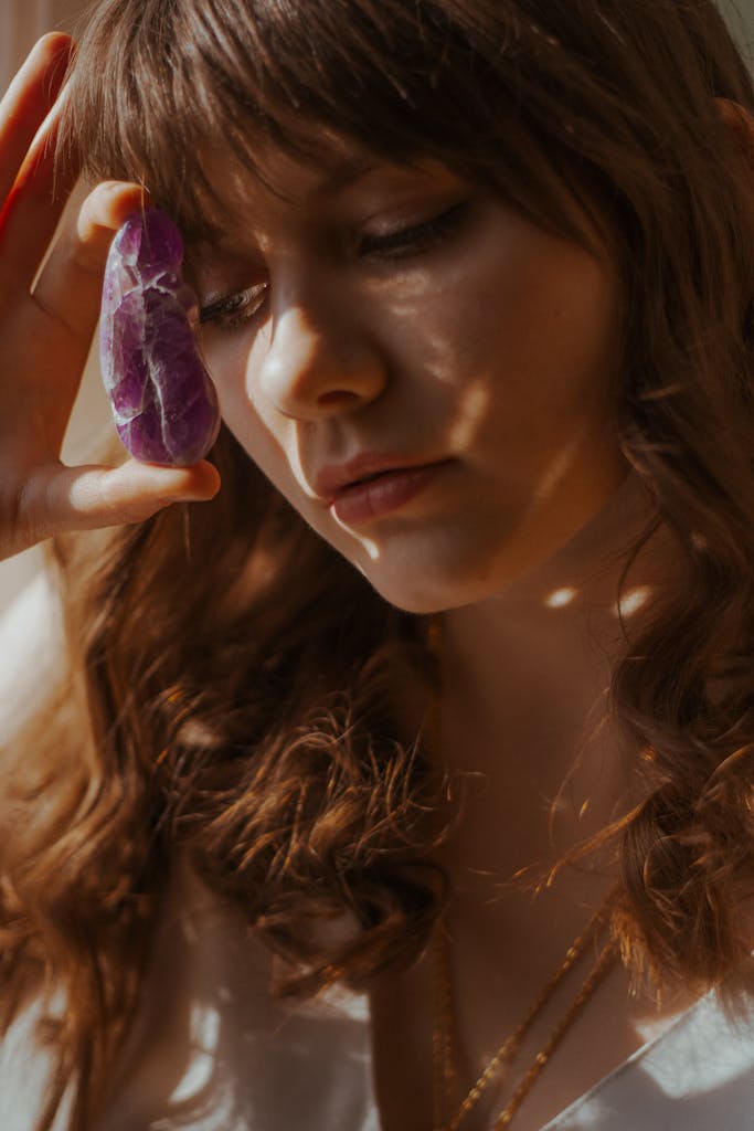 Calm female with long wavy hair standing in light room with natural amethyst in hand in bright sunlight at home