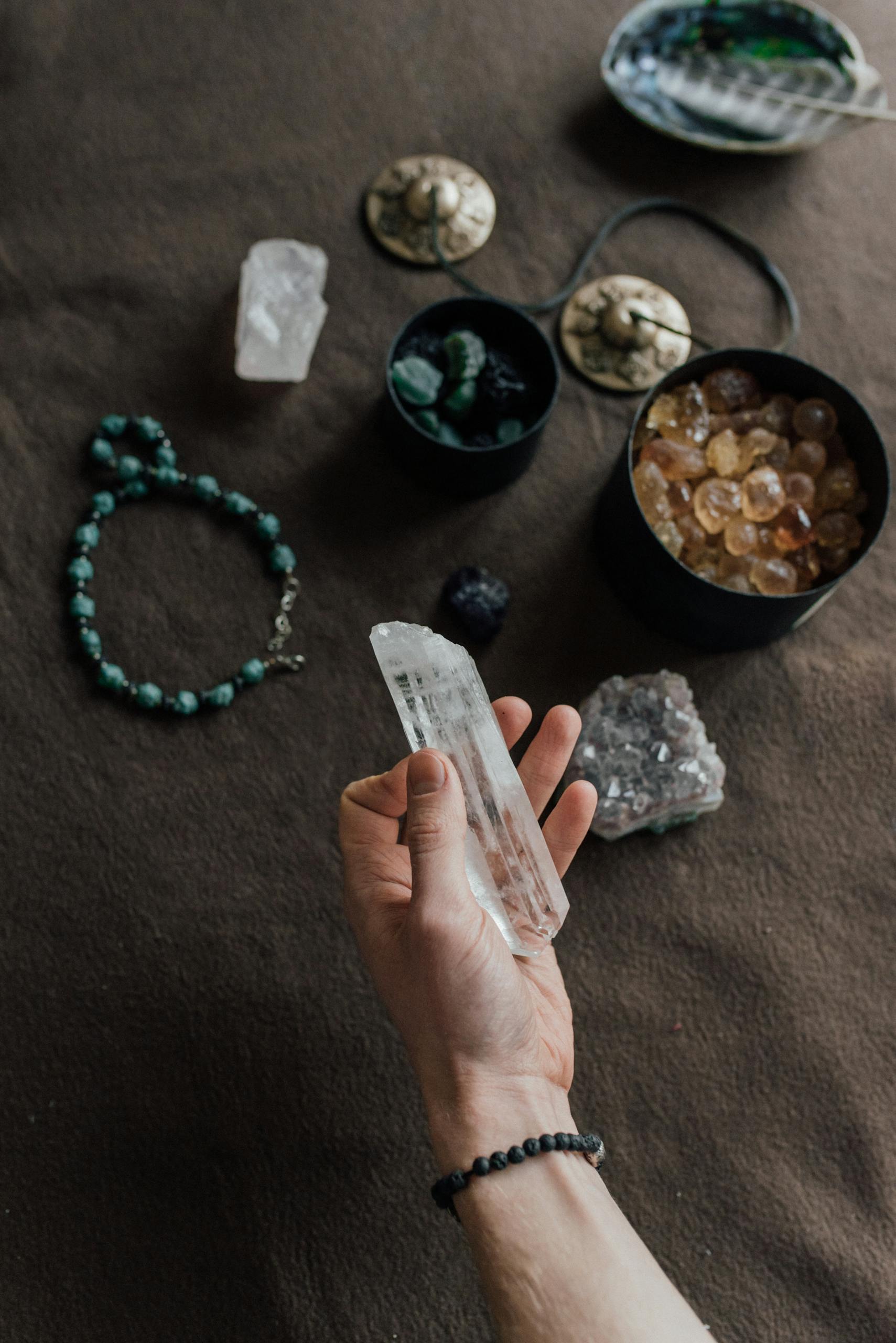 A hand holding a crystal with various stones and jewelry on a textured surface.